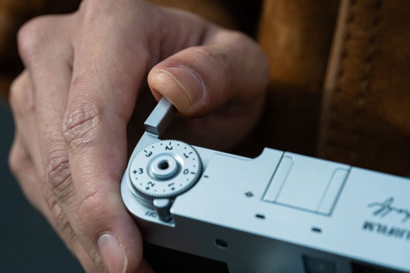 A close-up of a person’s hand adjusting the shutter speed dial on a silver camera, with part of their brown jacket visible in the background.
