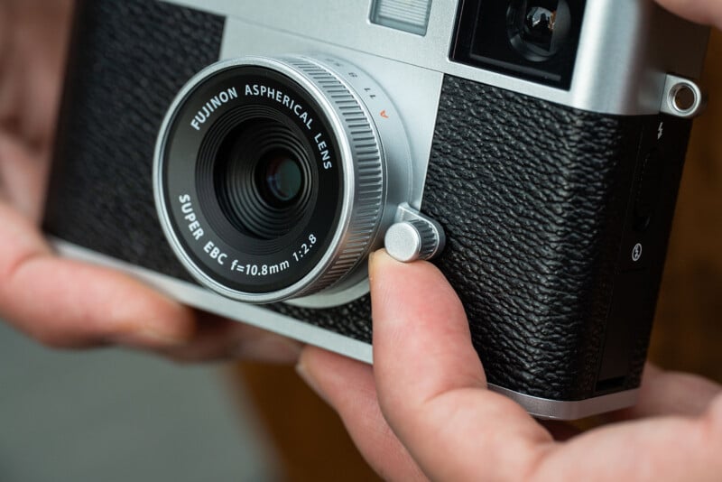 Close-up of hands holding a camera, focusing on the lens marked "Fujinon Aspherical Lens Super EBC f=10.8mm 1:2.8," with a textured black grip and silver detailing visible.