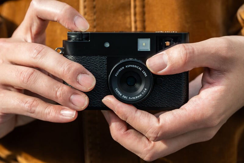 Close-up of two hands holding a black camera with a Fujinon aspherical lens, set against a blurred brown background. The person is wearing a brown jacket.