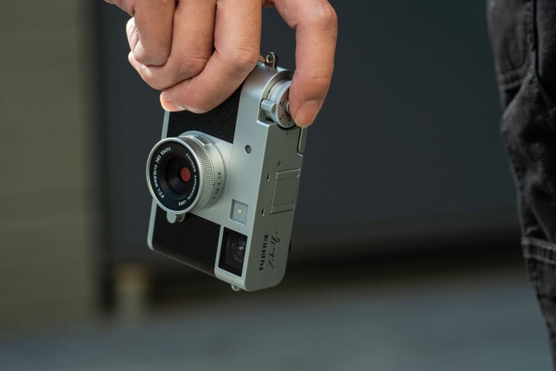 A hand holds a compact, silver and black Fujifilm X100-series camera by its top dial, with the camera hanging downward against a blurred background.
