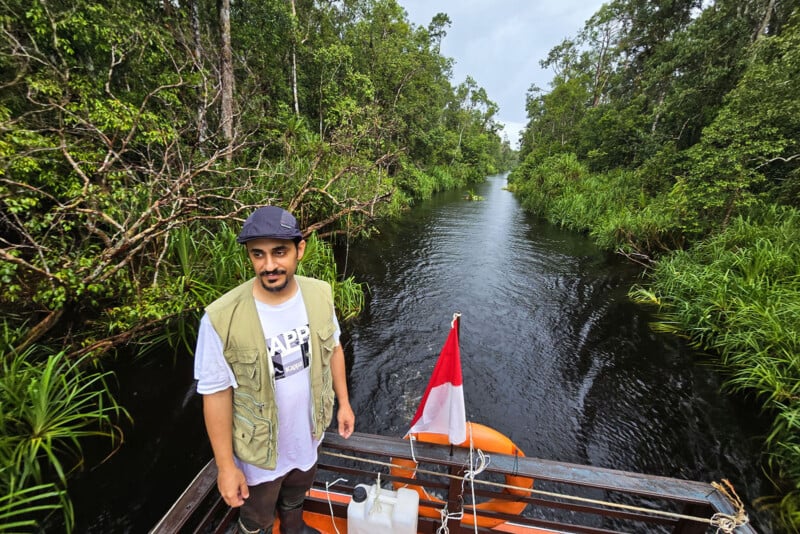 A man stands on a boat moving along a narrow jungle river, surrounded by dense green trees and vegetation. An Indonesian flag is visible at the back of the boat. The sky is overcast.