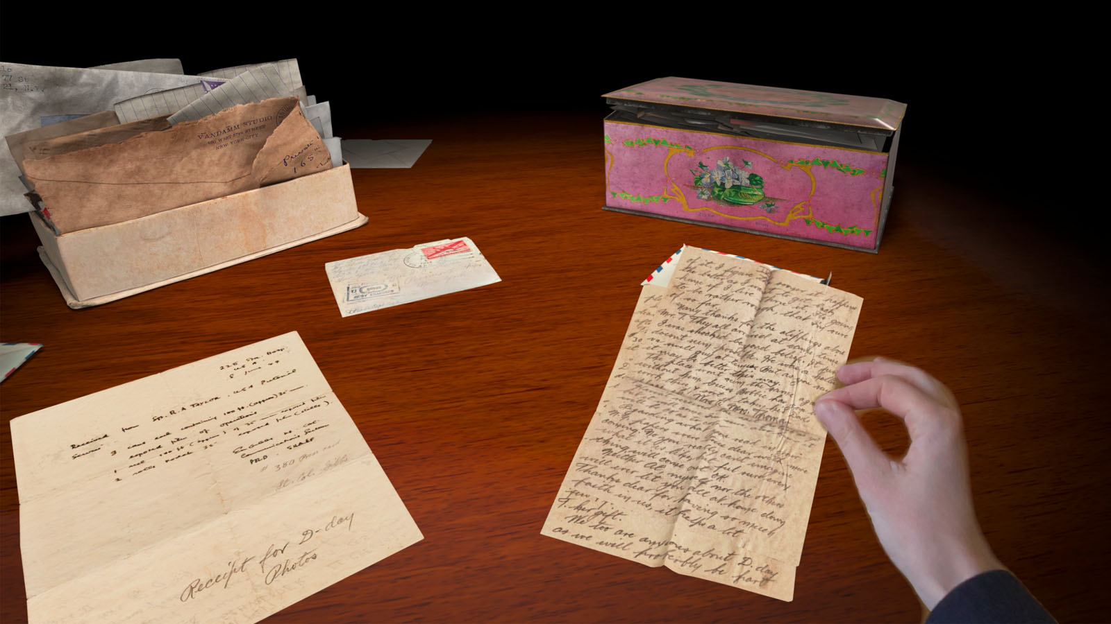 A hand holds an old handwritten letter over a wooden table, which also has more vintage letters, a typed document, and a decorative pink tin box.