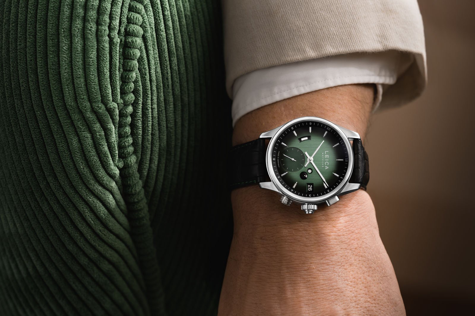 A person wearing a stylish green-faced wristwatch with a black band, dressed in a green corduroy garment and a beige shirt. The watch face features white accents and a date display.