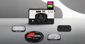 A retro-style camera with a pop-up flash and colored filters is displayed on a gray surface. Surrounding the camera are a close-up lens, a splitter lens, and a kaleidoscope lens attachment.