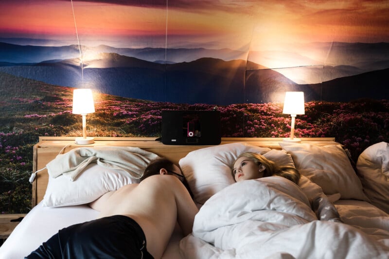 A shirtless person lies facedown in bed next to another person, who is awake and looking at them. Both are under white bedding, with two lit lamps on the headboard and a large scenic mountain mural in the background.