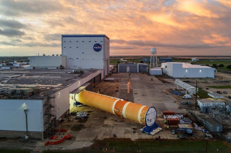 A large orange NASA rocket core stage is being wheeled out of a white building at sunset, surrounded by workers and equipment, with dramatic clouds and green fields in the background.