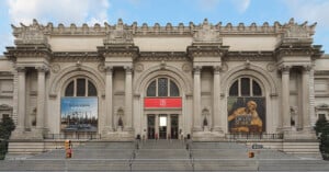 The grand entrance of the Metropolitan Museum of Art in New York City, featuring wide stone steps, arched doorways, columns, and large banners advertising exhibitions on either side of the main entrance.