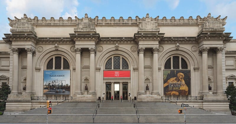 The grand entrance of the Metropolitan Museum of Art in New York City, featuring wide stone steps, arched doorways, columns, and large banners advertising exhibitions on either side of the main entrance.