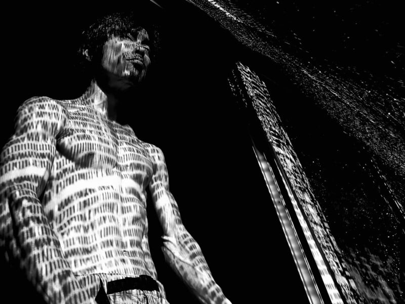 A shirtless person stands in dramatic lighting, with patterns of light and shadow resembling binary code or lines projected across their body and face, creating a striking and abstract visual effect.