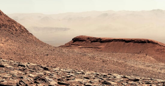 A rocky, reddish-brown Martian landscape with layered hills and mountains fades into a hazy, distant horizon under a pale sky. The terrain is dry and rugged, covered in loose rocks and dust.