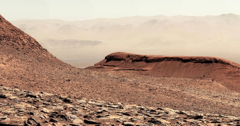 A rocky, reddish-brown Martian landscape with layered hills and mountains fades into a hazy, distant horizon under a pale sky. The terrain is dry and rugged, covered in loose rocks and dust.