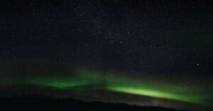 The night sky filled with stars and faint green aurora lights glowing above a dark, silhouetted landscape.