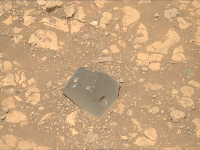 A translucent, irregularly shaped rock with smooth surfaces and a few small holes sits on a dusty, rocky, tan-colored ground. Scattered stones of various sizes surround the central rock.