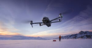 A large drone flies close to the camera over a snowy landscape at sunset; in the background, a person stands near a sled with mountains visible in the distance.