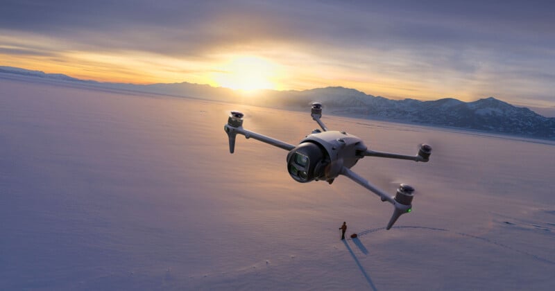 A large drone hovers over a snowy landscape at sunset, casting a shadow near a person standing on the snow with mountains visible in the background.