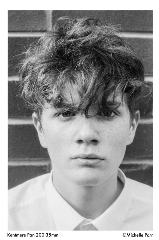 Black-and-white portrait of a young person with tousled hair, wearing a buttoned shirt, looking directly at the camera with a neutral expression. Brick wall in the background.