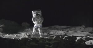 An astronaut in a spacesuit stands on a rocky, dusty lunar surface with scattered rocks and shadows, against the blackness of space.