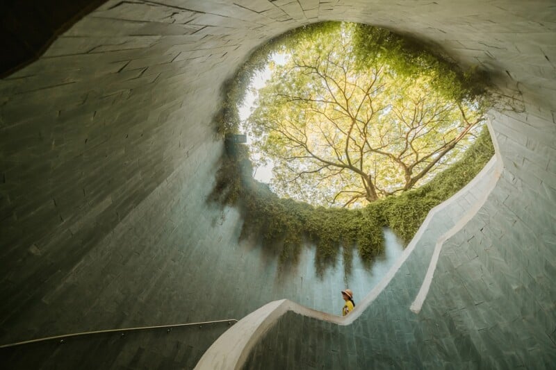 A person wearing a hat climbs a curved white staircase inside a circular stone tunnel with an opening above, revealing green trees and sunlight.