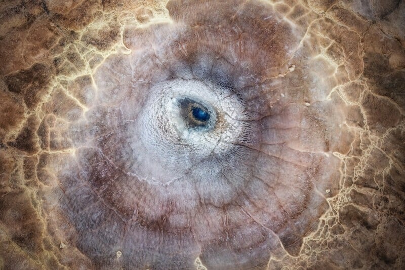 Aerial view of a circular, cracked, and textured surface resembling an eye, with brown, tan, and light beige tones radiating outward from a dark central spot. Patterns suggest dried earth or a geological formation.