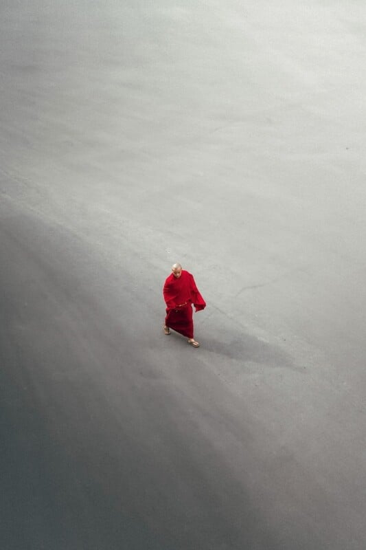 A person wearing a bright red robe walks alone on a vast, empty, gray surface, casting a faint shadow to the right. The scene is minimalist, with the person centered in the lower part of the image.