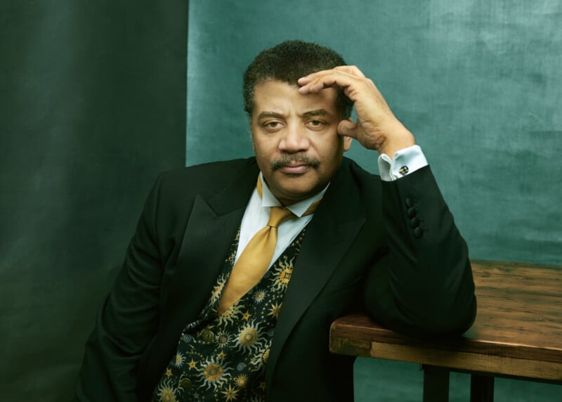 A man in a black suit with a patterned vest and gold tie sits at a wooden table, resting his head on his hand, looking thoughtfully at the camera against a green backdrop.
