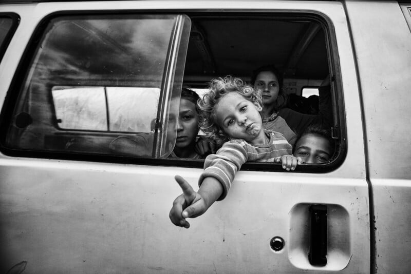 A group of children look out from the window of a van. One child with curly hair leans out, making a peace sign with their fingers. The image is in black and white.