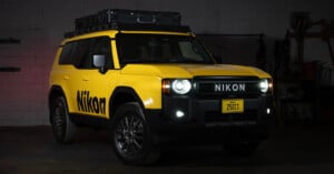 A yellow SUV with “Nikon” branding on the side and front, parked indoors. The vehicle has a roof storage box, black wheels, and a custom license plate reading “Z50II.” The headlights and fog lights are on.