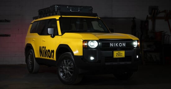 A yellow SUV with “Nikon” branding on the side and front, parked indoors. The vehicle has a roof storage box, black wheels, and a custom license plate reading “Z50II.” The headlights and fog lights are on.