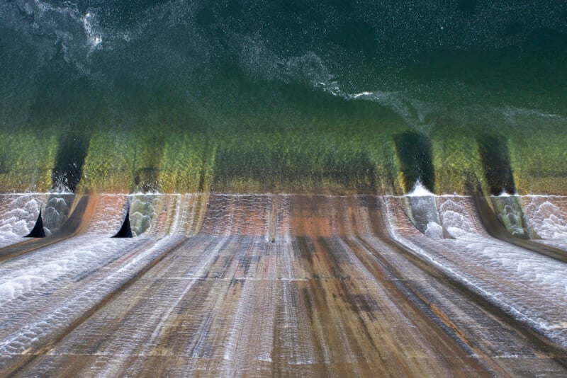 Aerial view of water flowing over the curved surface of a large dam spillway, creating white streaks and patterns as it cascades down into the structure below.