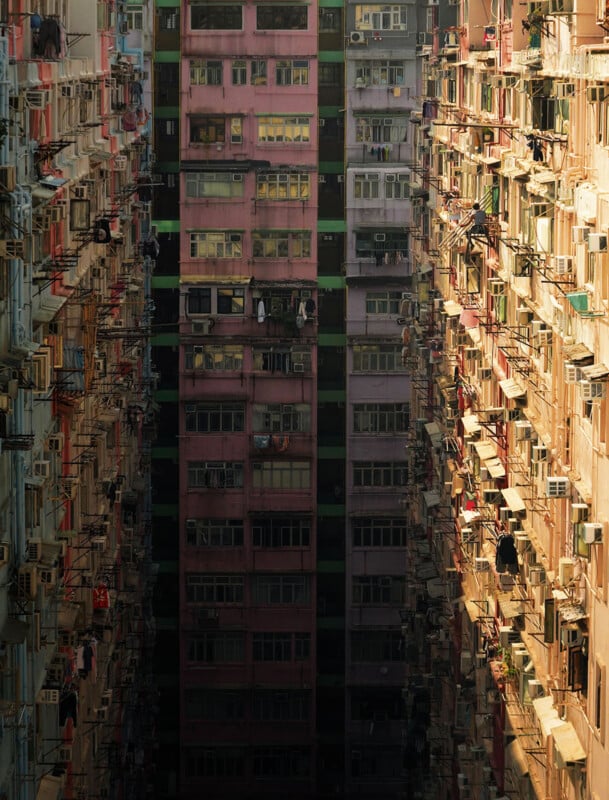 Tall, densely packed apartment buildings with numerous balconies and air conditioners face each other, creating a narrow alley with a dark, shadowy center. Warm sunlight illuminates the buildings on the right.