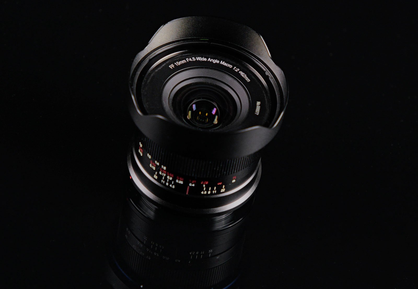 A wide-angle camera lens with a hood is placed on a glossy black surface, reflecting parts of the lens and its markings.