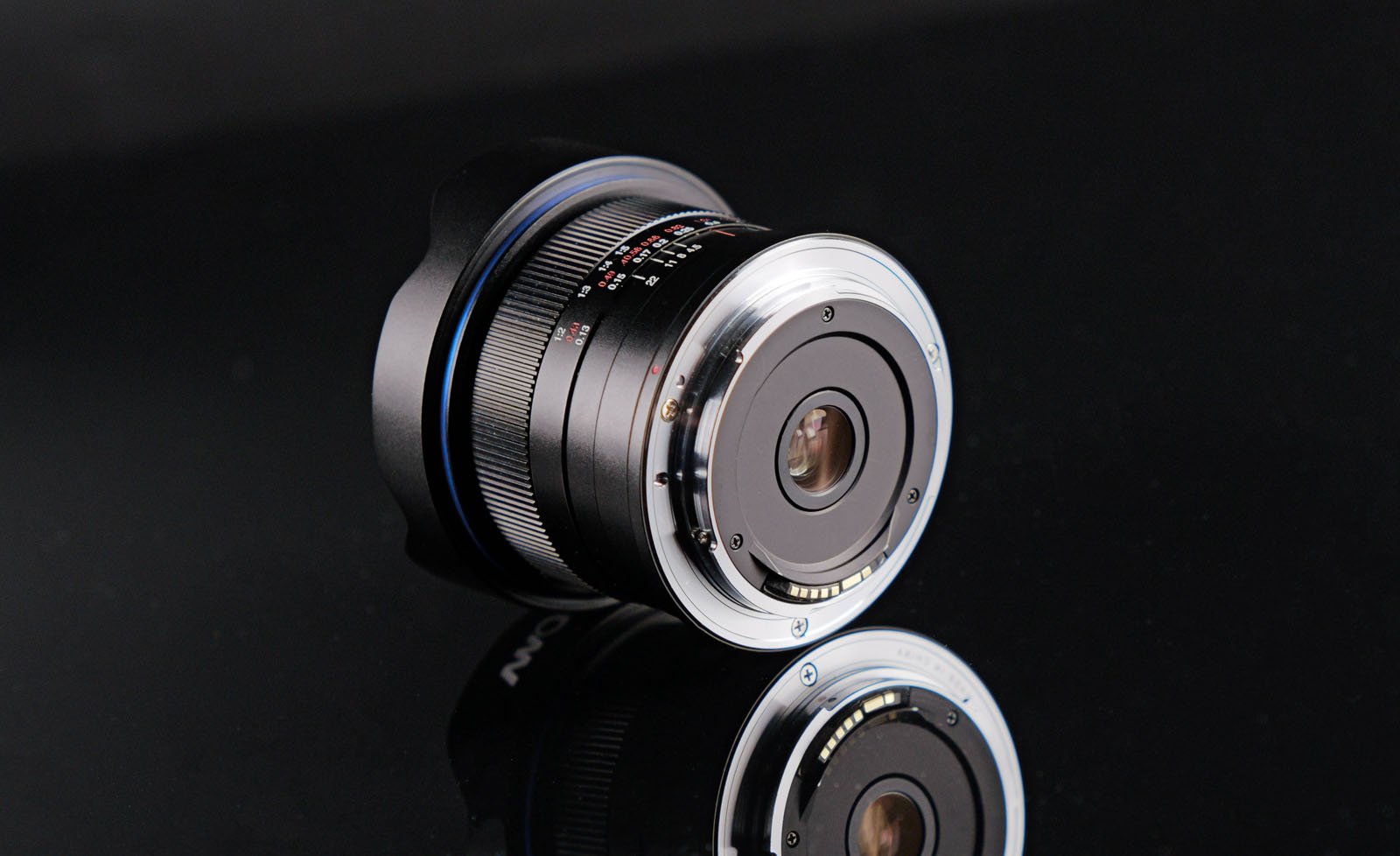 A camera lens with a silver metal mount and visible electronic contacts is lying on its side on a shiny black surface, reflecting the lens and its engraved markings.