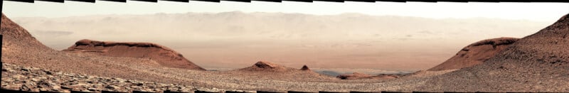 Wide panorama of a rocky, reddish Martian landscape with hills in the foreground and hazy mountains in the distance under a pale sky. The scene appears barren and desolate.