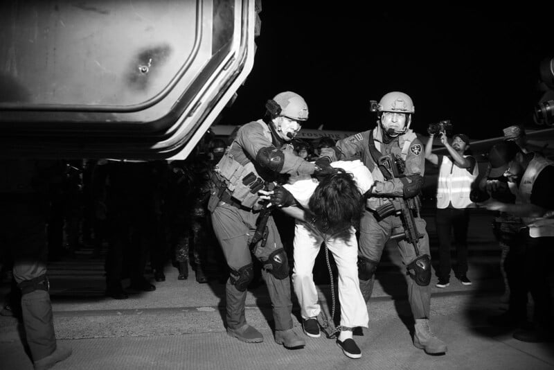 Two armed officers in tactical gear escort a person in white clothing with their head down. The scene is at night, with bright lights illuminating the group and additional people visible in the background.