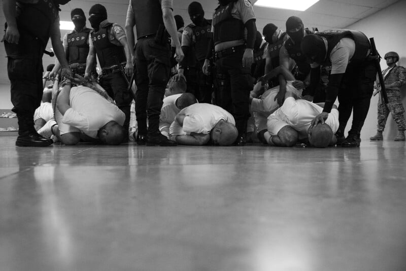 A group of handcuffed men in white shirts kneel on the floor with their heads down, surrounded by masked and armed officers standing over them in a brightly lit room.