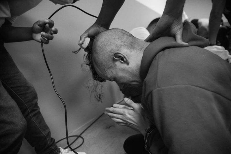 A person bows their head with hands pressed together as another shaves their head with electric clippers; others assist, creating an intimate and emotional scene in black and white.