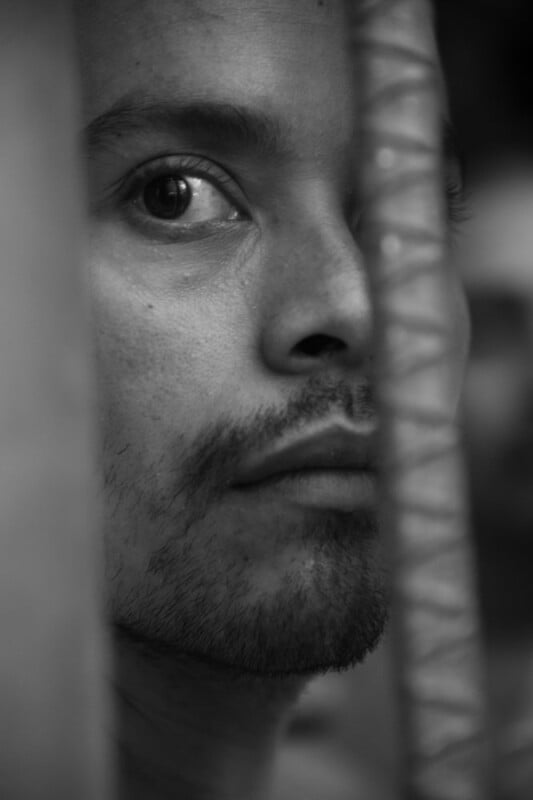 A black and white close-up photo of a man's face, partially obscured by a textured vertical bar. He has a serious expression, with visible stubble and intense eyes looking slightly upwards.