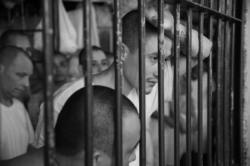 A group of men stand closely together behind metal bars in a dimly lit room. One man grips the bars and looks out pensively, while others appear somber and reflective in the background.