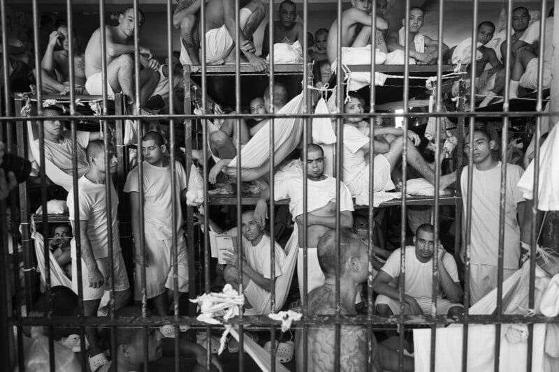 A crowded prison cell filled with men wearing light-colored clothing, some standing, sitting, or lying on hammocks and bunks, behind metal bars. The cell appears cramped and overcrowded.