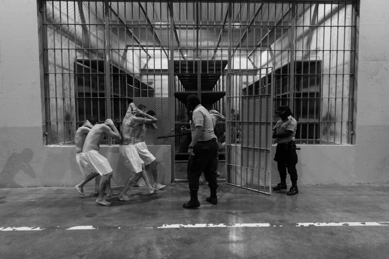 Three shirtless prisoners in white shorts walk with their hands on their heads, escorted by two armed guards in a barred prison corridor. The scene is harshly lit and the mood is tense.