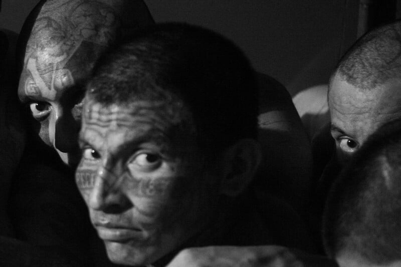 Three men with tattooed faces sit closely together in dim light, looking towards the camera. The image is in black and white, highlighting the intensity in their expressions and the details of their tattoos.