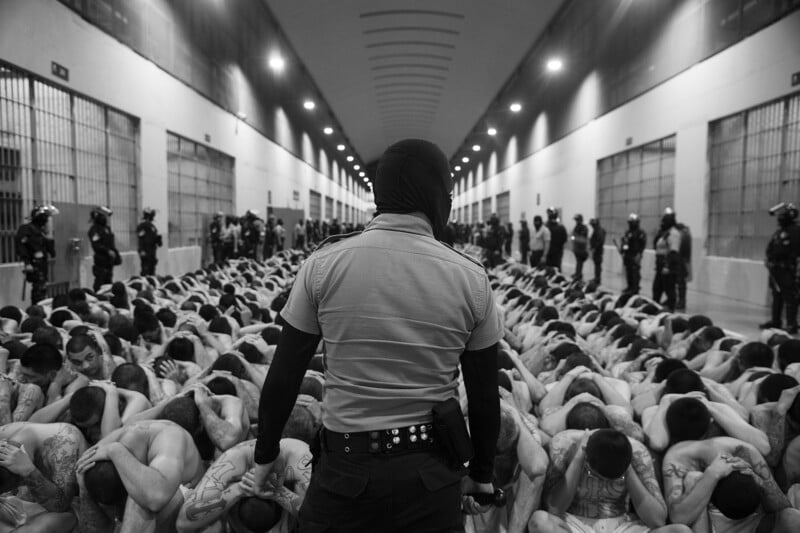 A masked officer stands facing a large group of shirtless prisoners sitting closely together on the floor of a prison hallway, hands on their heads, with armed guards lined along the corridor walls.