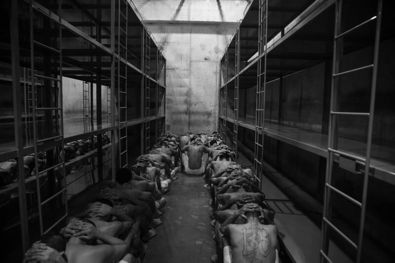 A group of shirtless men sit closely together on the floor of a stark room between empty metal bunk beds, facing away with their hands on their heads under harsh lighting.