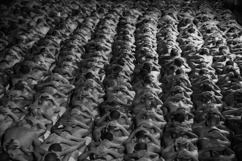A large group of people sit close together in uniform rows, all facing the same direction, with their hands clasped behind their heads. The image is in black and white.