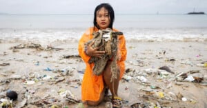 A woman in an orange raincoat kneels on a beach holding a large pile of trash, surrounded by litter and plastic debris, with the ocean and cloudy sky in the background.