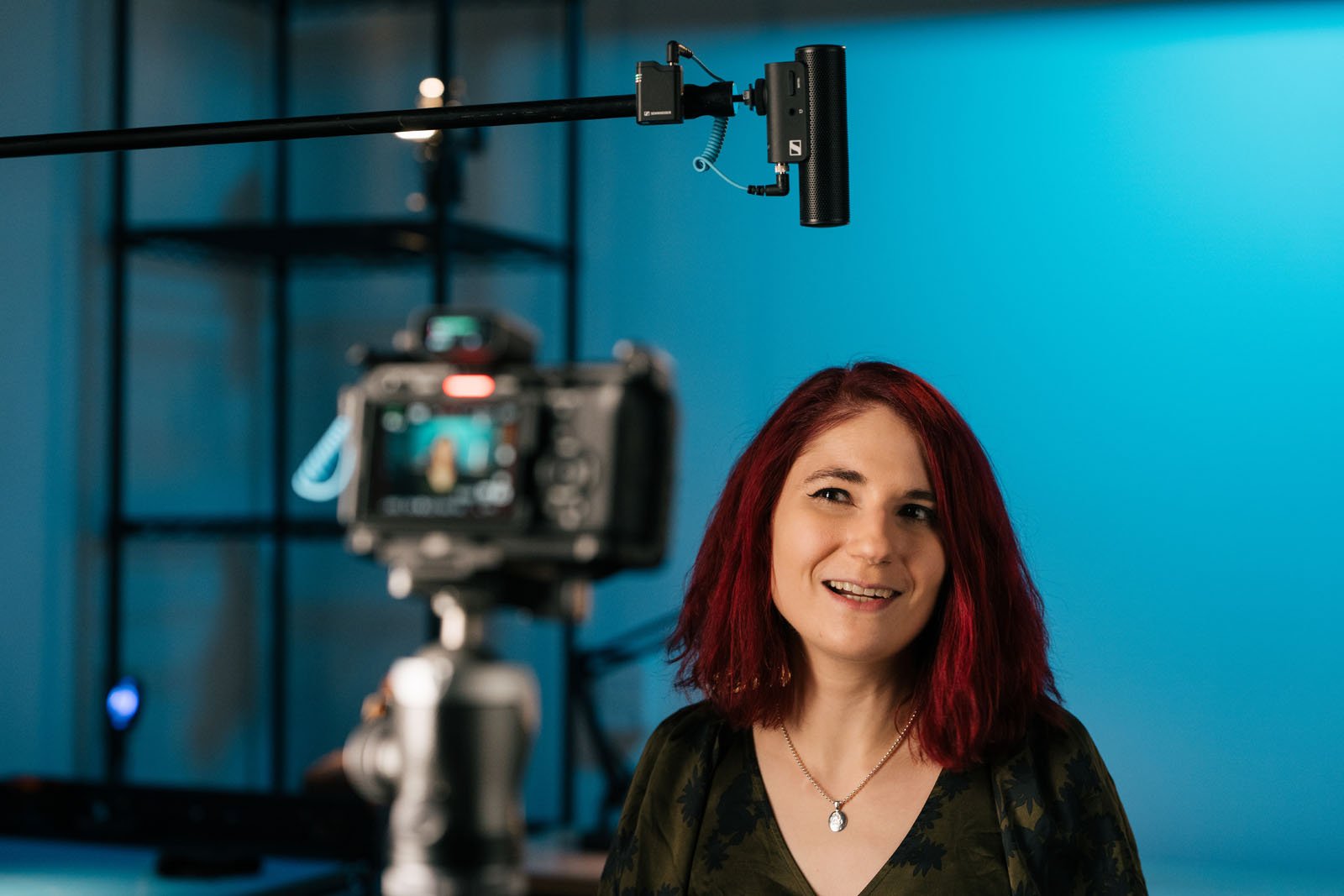 A woman with red hair smiles while standing in front of a camera and microphone in a studio with blue lighting. Shelves and equipment are visible in the background.
