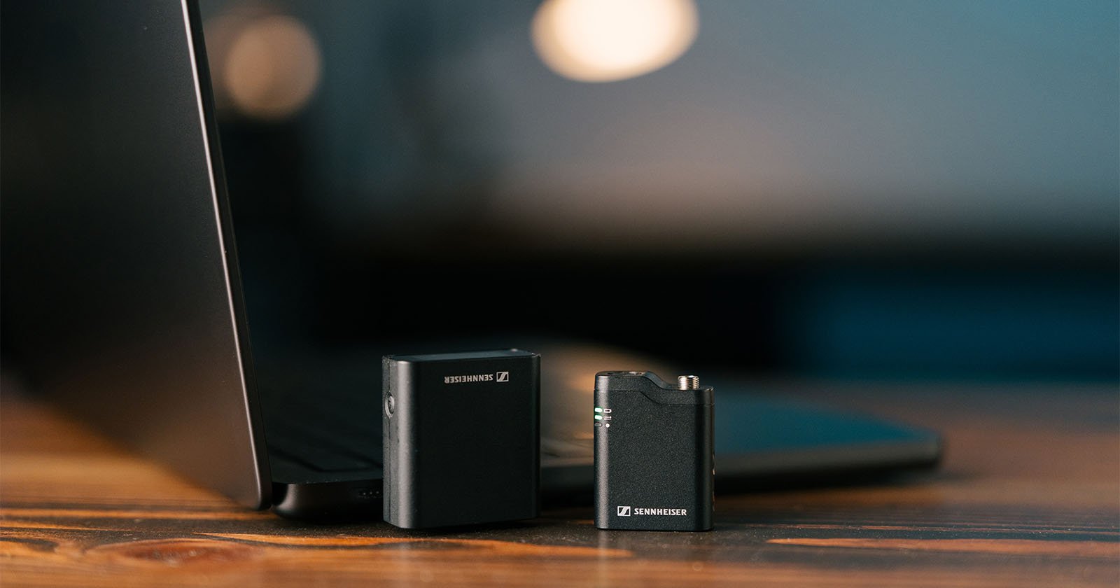 Two Sennheiser audio transmitters sit on a wooden desk next to a partially open black laptop, with a blurred background featuring warm lighting.
