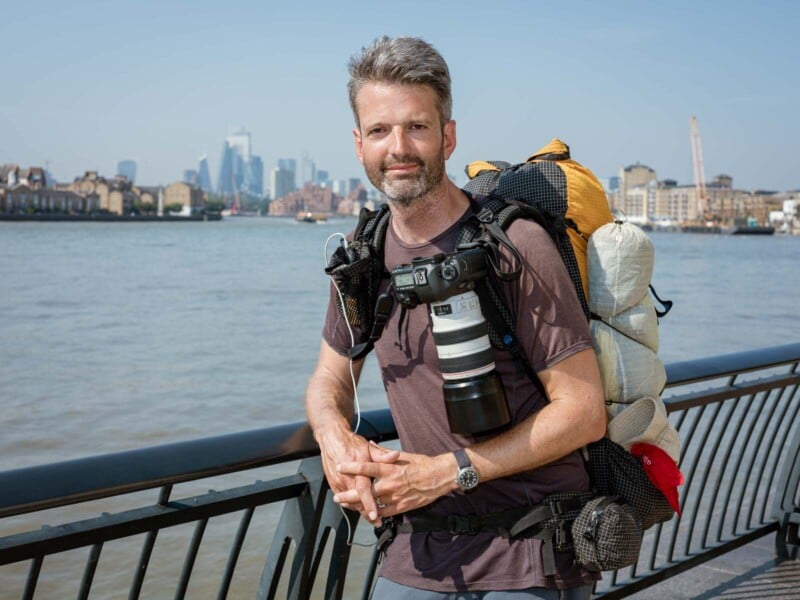 A man with graying hair and beard stands by a riverfront railing, wearing a backpack with gear and a camera hanging from his neck. City buildings and a blue sky are visible across the water in the background.