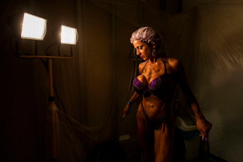A female bodybuilder in a purple bikini and shower cap stands under bright lights in a backstage area with plastic sheets and shadows, highlighting her muscles and focused expression.
