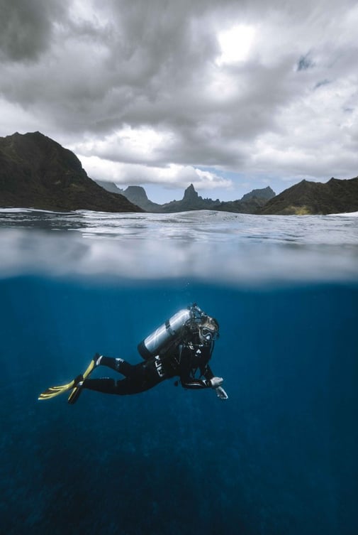 A scuba diver swims underwater in clear blue ocean, wearing fins and scuba gear. Above the water, rugged mountains and a cloudy sky are visible in the background.